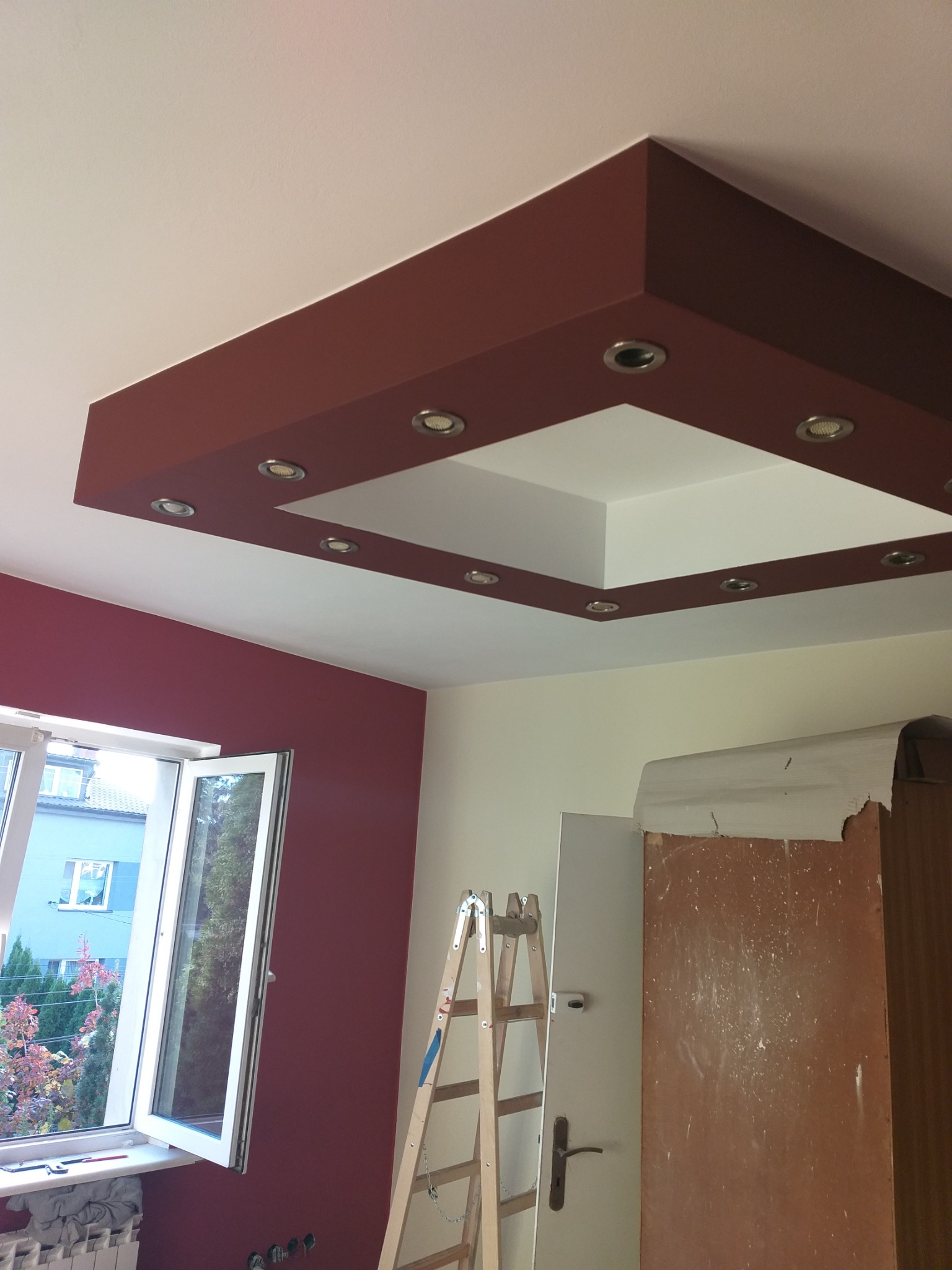 Pomieszczenie w trakcie remontu z pomalowaną na bordowo ścianą i sufitem podwieszanym z oświetleniem halogenowym, widoczna drabina i okno z widokiem na zewnątrz.