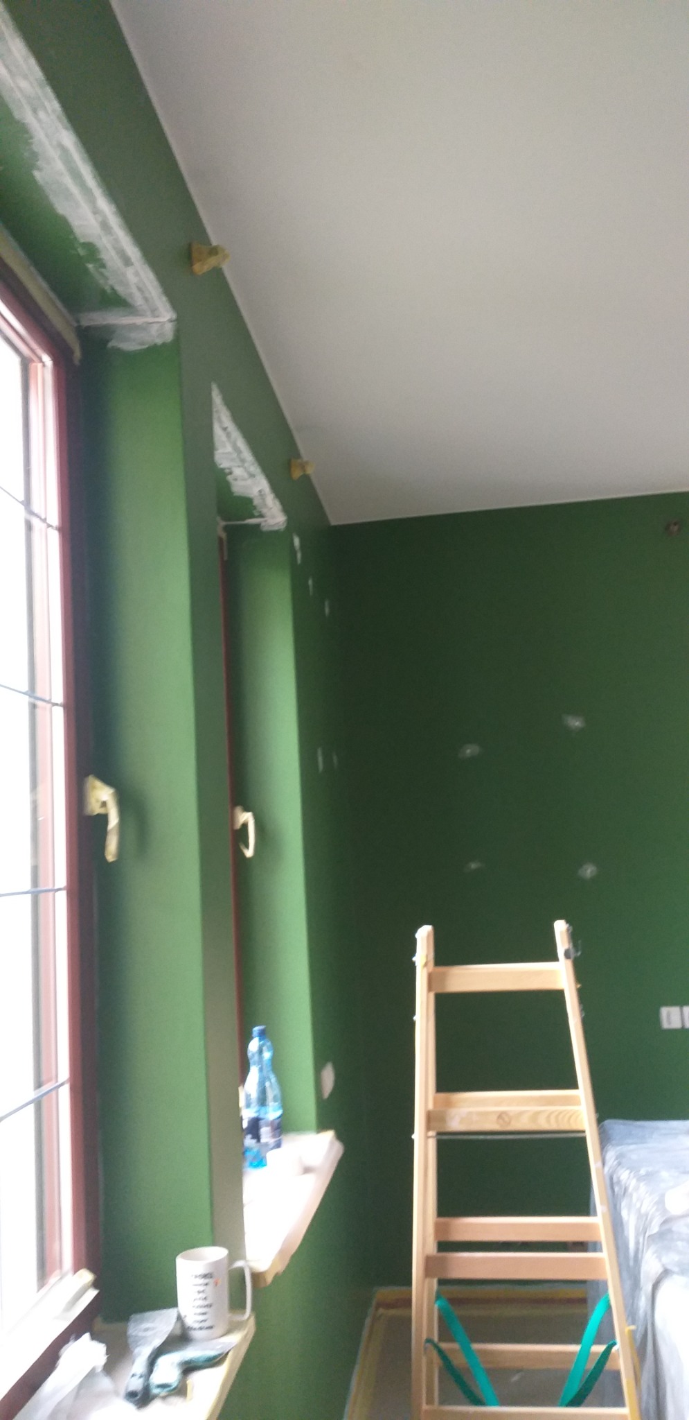 Wnętrze pokoju w trakcie malowania na zielono, widoczne ślady szpachli przy suficie i oknie, drabina oparta o ścianę, butelka wody i kubek na parapecie.