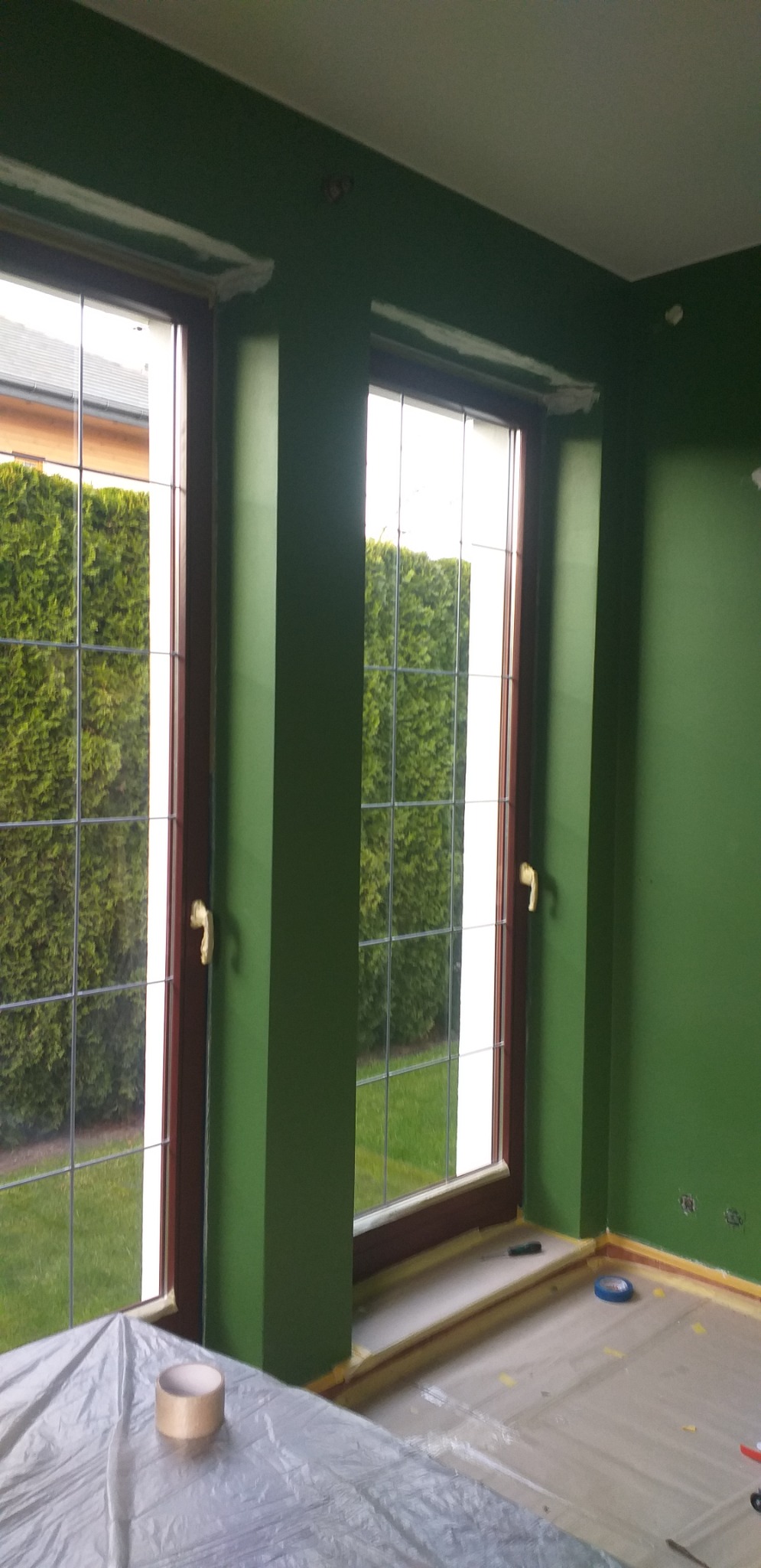Wnętrze pokoju po malowaniu na zielono, z widocznymi dwoma oknami i zabezpieczoną folią podłogą; ślady taśmy malarskiej na framugach okiennych i przy podłodze.