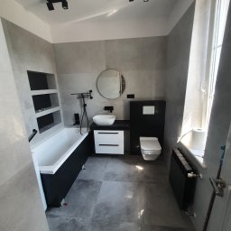 Piter-Bud Piotr Papiernik - Nowoczesna łazienka w szarościach z czarnymi akcentami: wanna z półkami, umywalka z szafką, toaleta i grzejnik. Minimalistyczne oświetlenie punktowe.