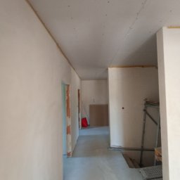 Piter-Bud Piotr Papiernik - Wnętrze w trakcie szpachlowania: gładkie, jasne ściany i sufit z widocznymi łączeniami płyt gipsowych. W tle widoczne rusztowanie i elementy wyposażenia.