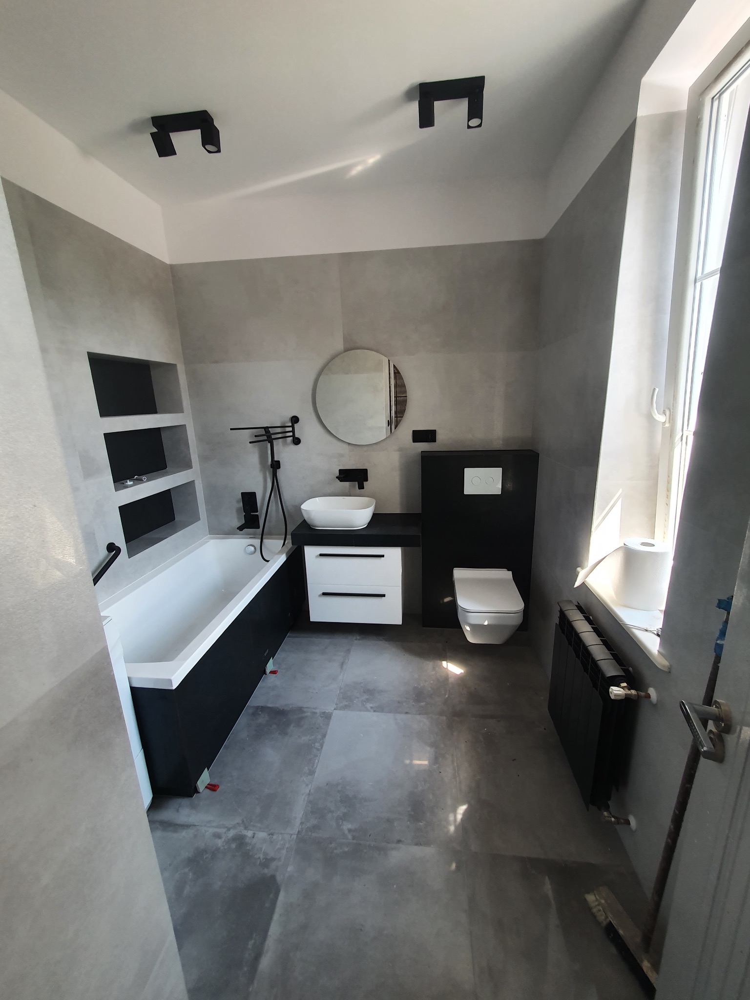 Nowoczesna łazienka w szarościach z czarnymi akcentami: wanna z półkami, umywalka z szafką, toaleta i grzejnik. Minimalistyczne oświetlenie punktowe.