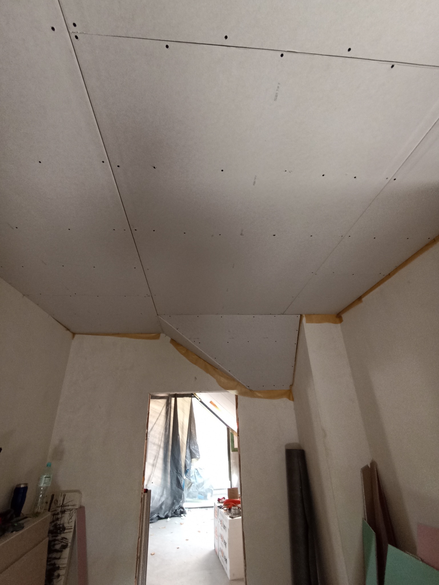 Montaż płyt gipsowo-kartonowych na suficie poddasza, widoczne łączenia i mocowania. Prace wykończeniowe w trakcie realizacji, widok na wnętrze przez otwarte przejście.