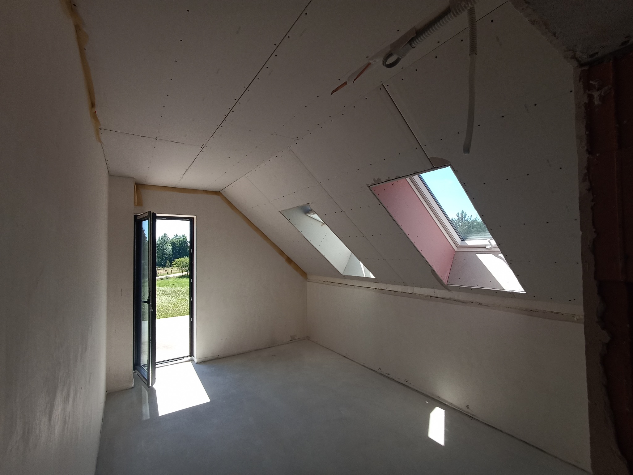 Poddasze z oknami dachowymi w trakcie wykańczania: ściany przygotowane do szpachlowania, widoczne płyty gipsowo-kartonowe i otwarte drzwi balkonowe z widokiem na zieleń.