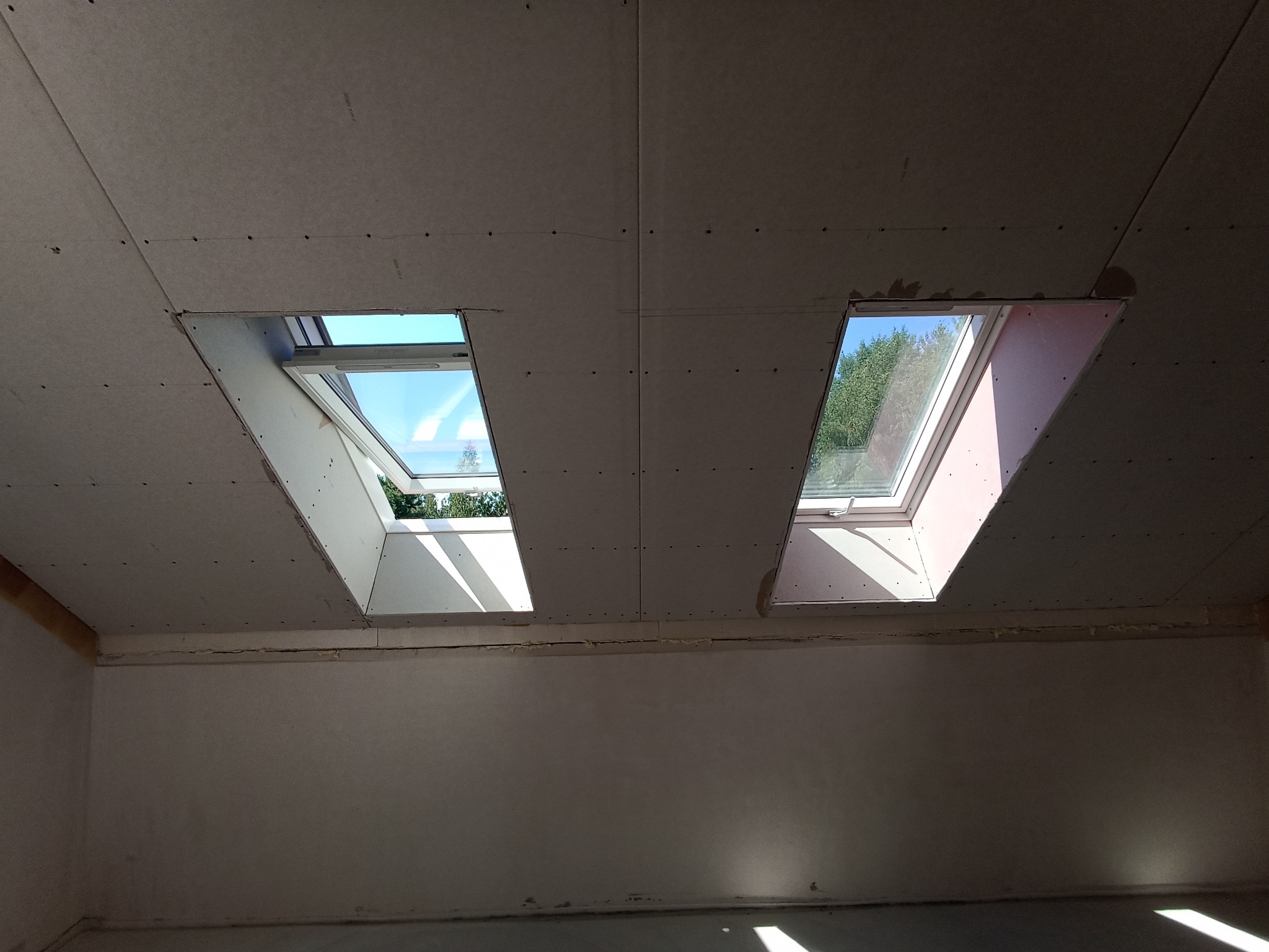 Płyty gipsowo-kartonowe na poddaszu z dwoma oknami dachowymi w trakcie montażu. Widoczne łączenia płyt i otwory na wkręty. Częściowo oświetlone wnętrze.