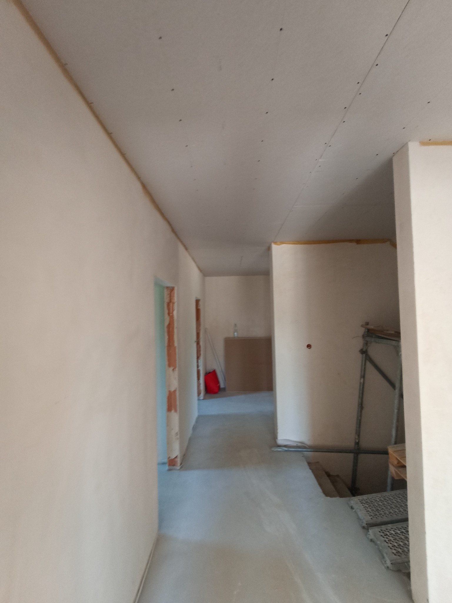 Wnętrze w trakcie szpachlowania: gładkie, jasne ściany i sufit z widocznymi łączeniami płyt gipsowych. W tle widoczne rusztowanie i elementy wyposażenia.