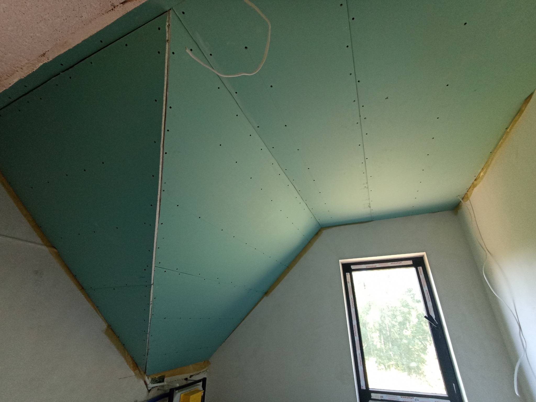 Poddasze z oknem dachowym, wykończone płytami gipsowo-kartonowymi w kolorze zielonym, widoczne łączenia i wkręty. Ocieplenie widoczne w szczelinach.
