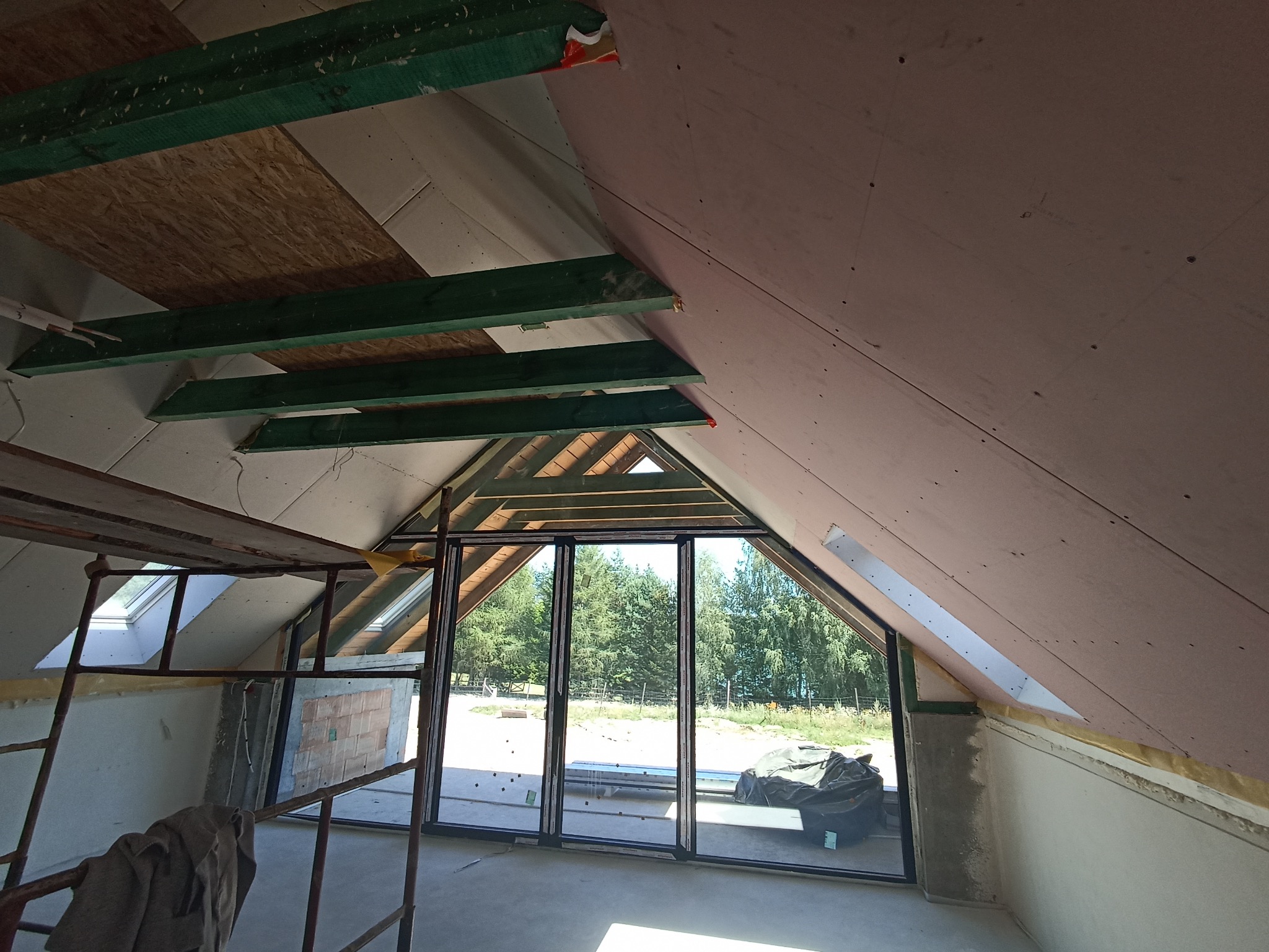 Poddasze w trakcie ocieplania z widoczną konstrukcją dachu, oknami i rusztowaniem. Ściany wykończone płytami gipsowo-kartonowymi, widoczne elementy izolacji.