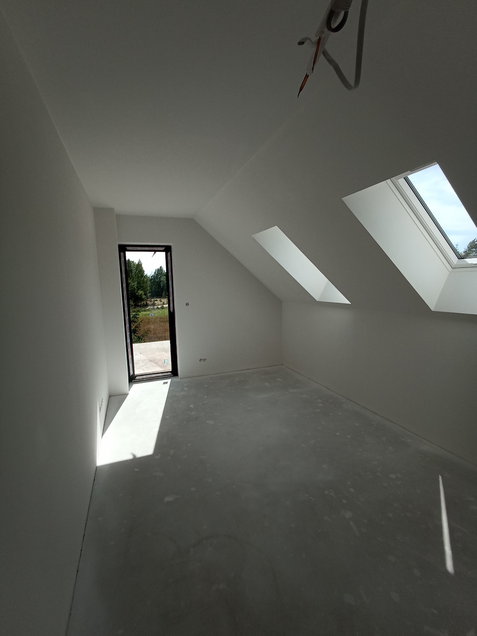 Jasne poddasze po szpachlowaniu i malowaniu na biało, z oknami dachowymi i otwartym wyjściem na zewnątrz. Widoczne ślady po pracach na podłodze.