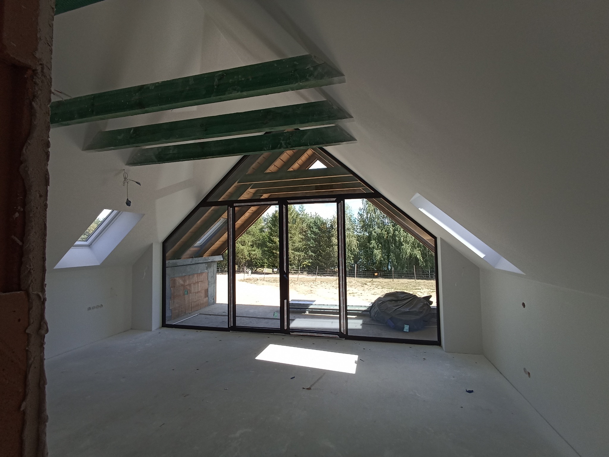 Poddasze z oknami dachowymi i dużym oknem balkonowym z widokiem na zieleń. Ściany pomalowane na biało, widoczne elementy konstrukcyjne dachu i fragment ściany w stanie surowym.