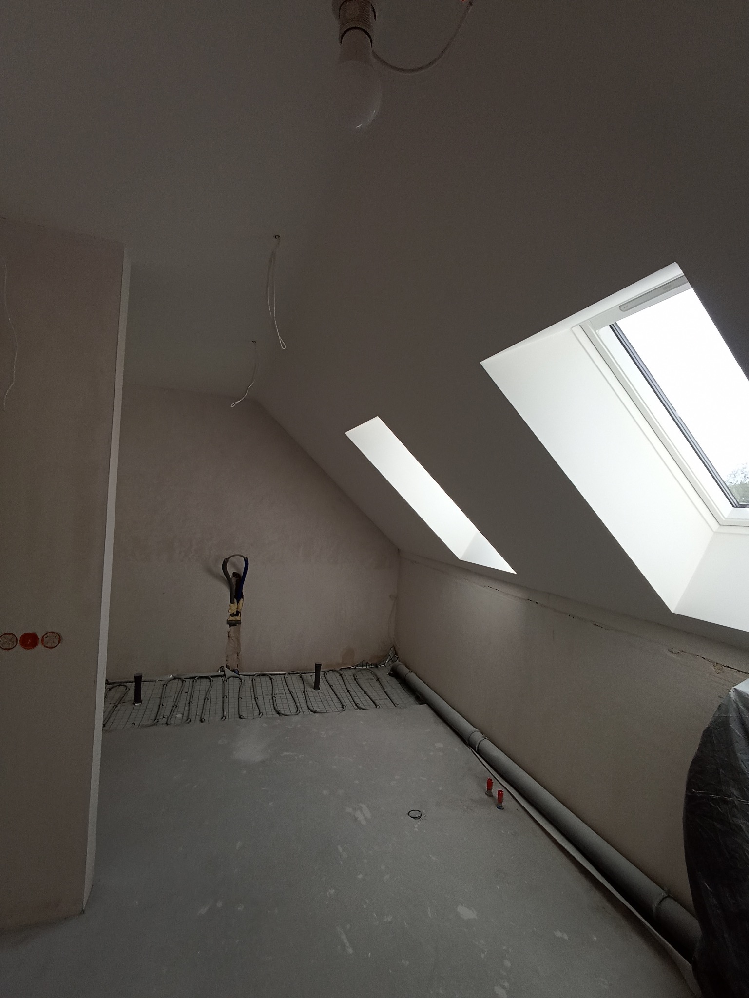 Poddasze w Mrągowie z oknami dachowymi i instalacją ogrzewania podłogowego przed wylewką. Widoczne przewody elektryczne i rury. Ściany przygotowane do dalszych prac.