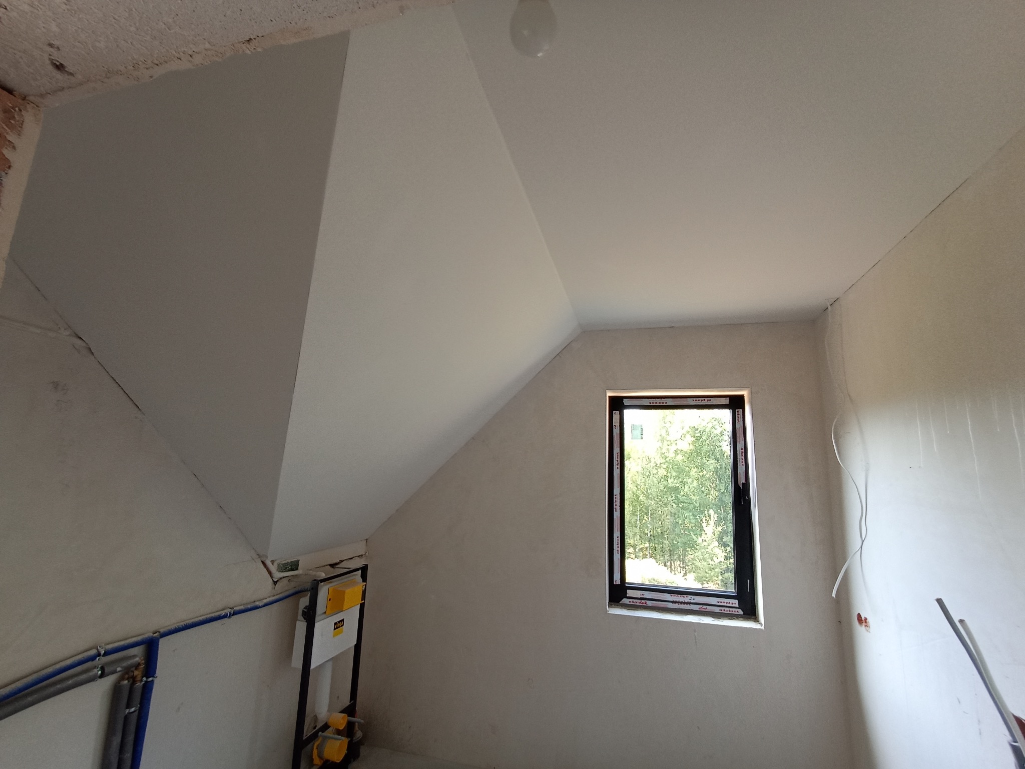 Wnętrze poddasza po szpachlowaniu i malowaniu na biało, z oknem w czarnej ramie. Widoczne instalacje hydrauliczne i elektryczne przed wykończeniem.