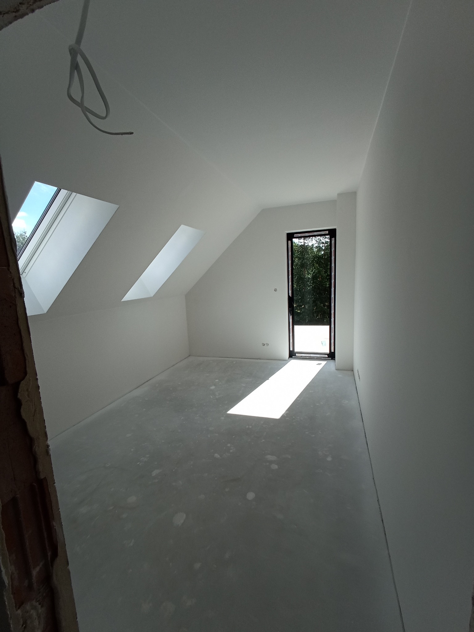 Jasne poddasze po szpachlowaniu i malowaniu na biało, z dwoma oknami dachowymi i czarnymi drzwiami balkonowymi. Na podłodze widoczne ślady po pracach wykończeniowych.