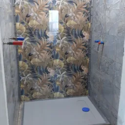 Wykończona kabina prysznicowa z zamontowanym brodzikiem i ozdobnymi płytkami ściennymi w kwiatowe wzory oraz marmurkowymi płytkami po bokach, widoczne przyłącza wodne.