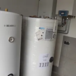 Dwa białe zbiorniki na wodę, jeden marki Biawar z widocznym manometrem, drugi marki Kospel owinięty folią, w tle biały piec gazowy i szara ściana wyłożona płytkami, widoczne elementy instalacji...