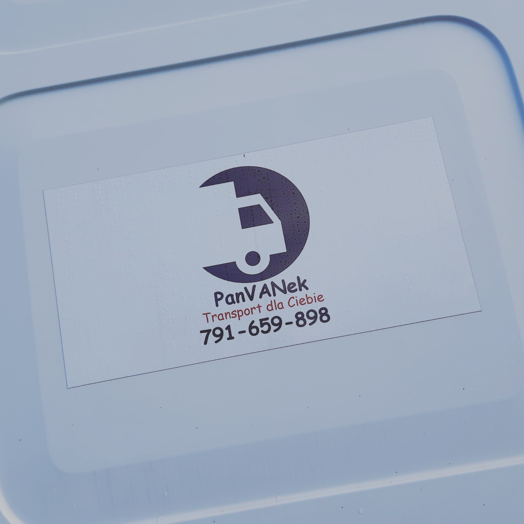 Naklejka z logo firmy transportowej 'PanVANek Transport dla Ciebie' z numerem telefonu na białej powierzchni, pokrytej kroplami wody.
