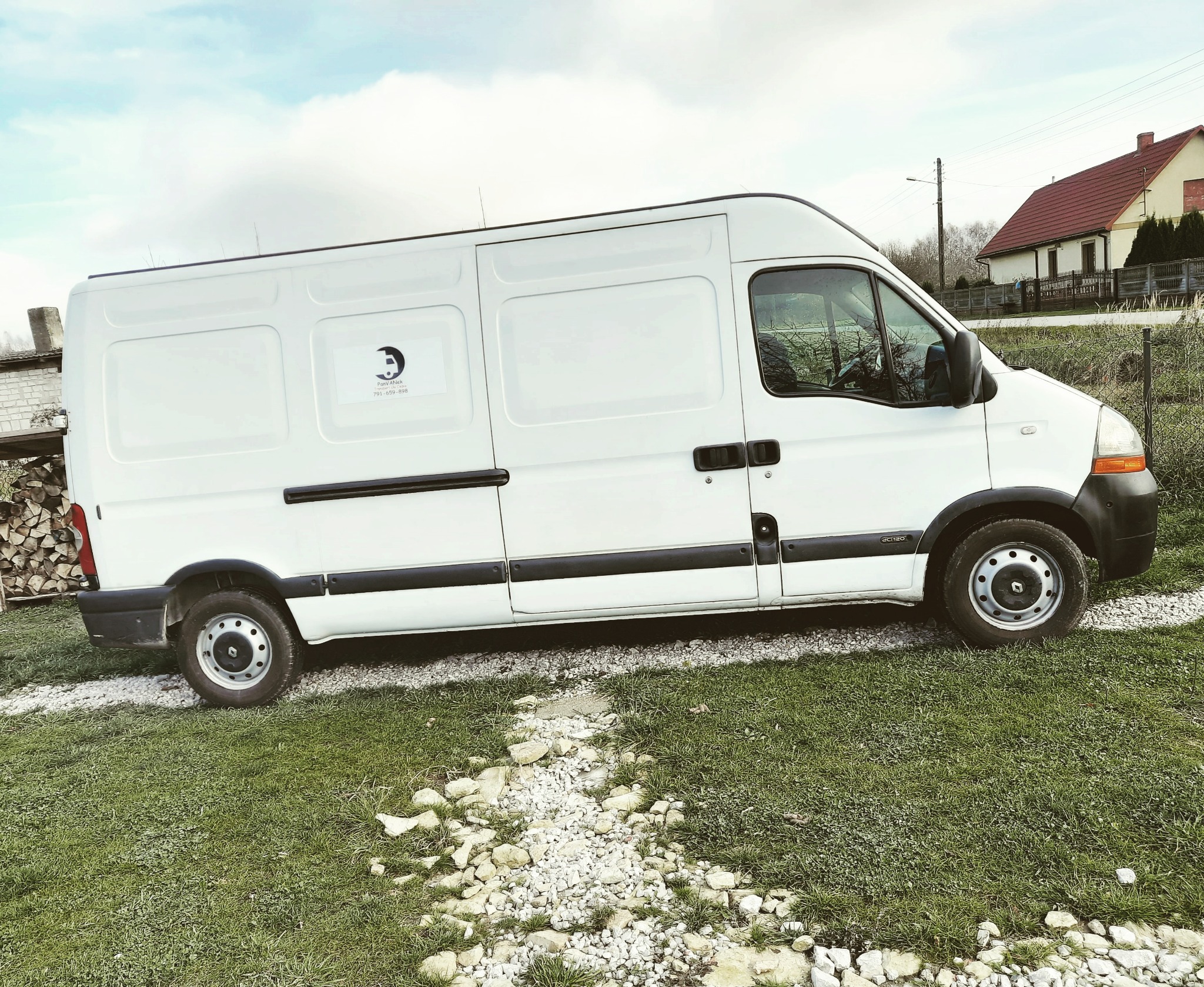 Biały Renault Master zaparkowany na trawie z widocznym logo firmy na boku, w tle budynek z czerwonym dachem i sterta drewna.