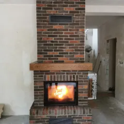 Murowany kominek z cegły klinkierowej z drewnianą półką i wkładem kominkowym, widoczne detale wykończenia wnętrza w trakcie realizacji.