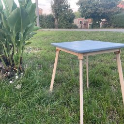 Ryweko - Niski, kwadratowy stolik z pomalowanym na niebiesko blatem i surowymi drewnianymi nogami stoi na trawie, obok kępy zielonych roślin. W tle widoczny fragment budynku i drzewa.