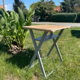 Składany, drewniany stolik z szarą metalową konstrukcją, ustawiony na trawie obok kępy zielonych roślin.