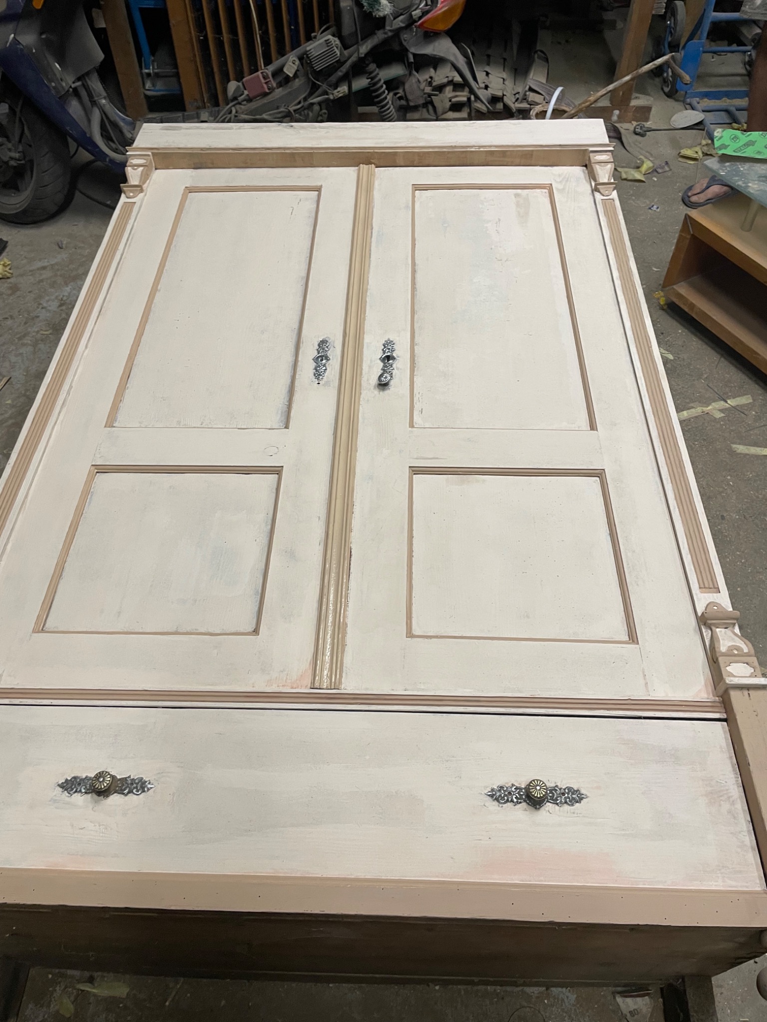 Odrestaurowana szafa dwudrzwiowa z pojedynczą szufladą, pomalowana na kremowo z ozdobnymi uchwytami, widoczna w warsztacie z motorem w tle.