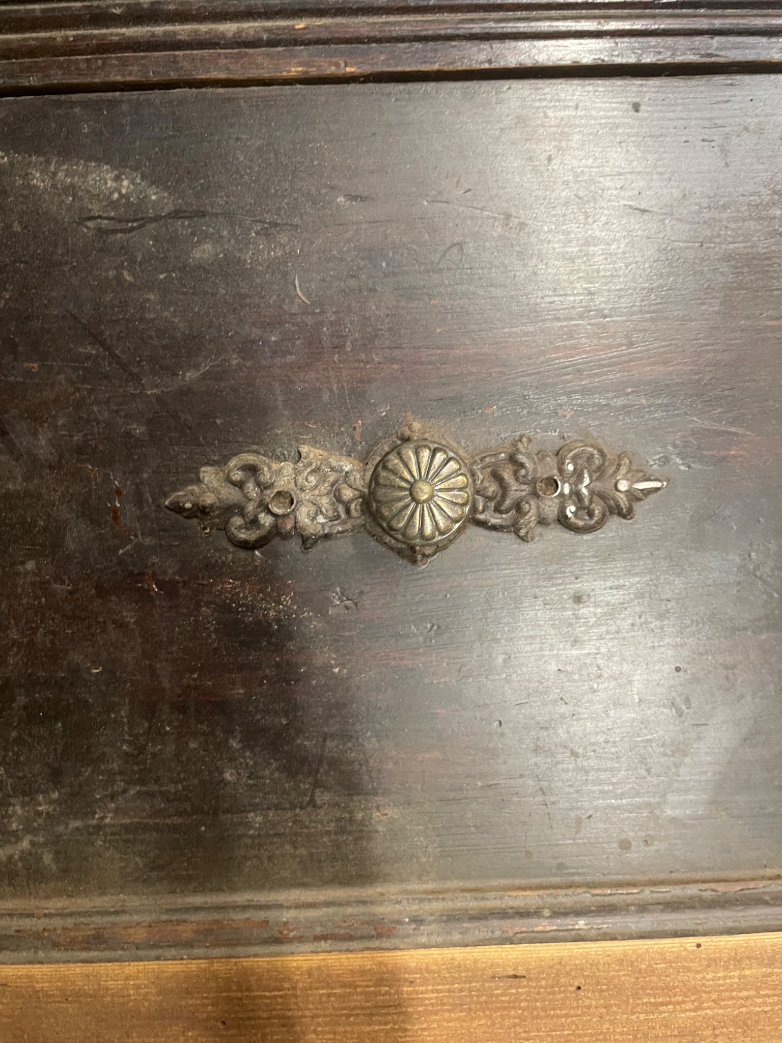 Fragment starej, ciemnobrązowej drewnianej szuflady z metalowym uchwytem w kształcie stylizowanej rozety z ornamentami.