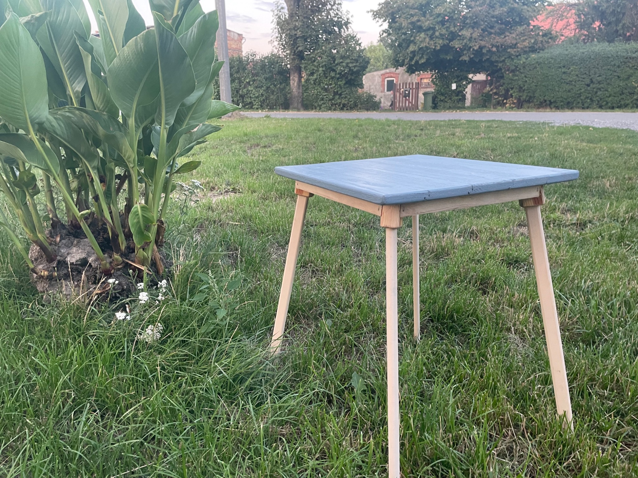Niski, kwadratowy stolik z pomalowanym na niebiesko blatem i surowymi drewnianymi nogami stoi na trawie, obok kępy zielonych roślin. W tle widoczny fragment budynku i drzewa.