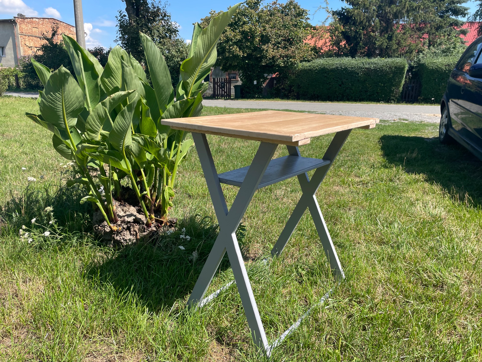 Składany, drewniany stolik z szarą metalową konstrukcją, ustawiony na trawie obok kępy zielonych roślin.