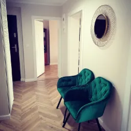 Dwa szmaragdowe, welurowe krzesła na czarnych nóżkach w przedpokoju z parkietem ułożonym we wzór jodełki, okrągłe lustro w złotej ramie na ścianie, otwarte drzwi w tle.