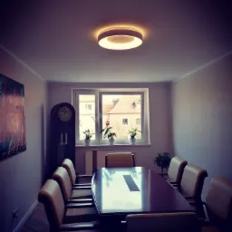 Sala konferencyjna z dużym, lakierowanym stołem, krzesłami z beżową tapicerką, wysokim zegarem stojącym i obrazem na ścianie, oświetlona okrągłą lampą sufitową. W tle widoczne okno z roślinami.