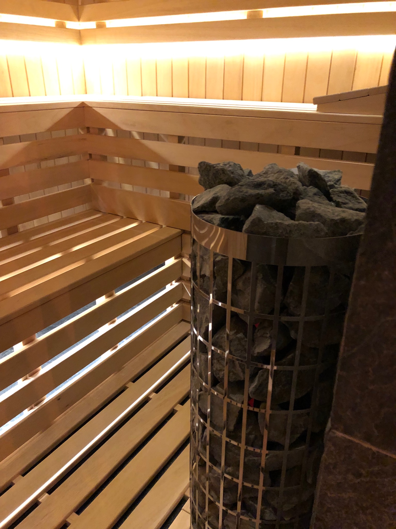 Wnętrze sauny z drewnianymi ławkami i piecem wypełnionym kamieniami, oświetlone ciepłym światłem.
