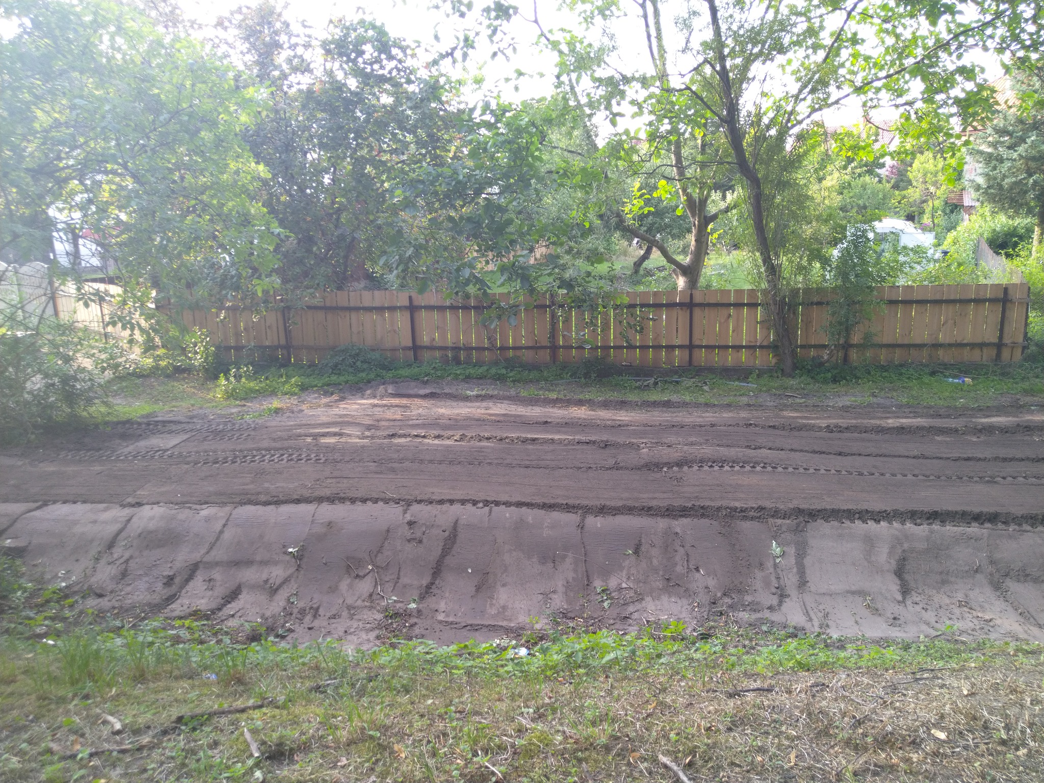 Przygotowany teren pod budowę z widocznymi śladami opon, ogrodzony drewnianym płotem, w tle zieleń drzew