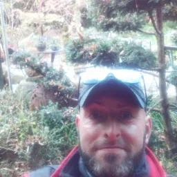 Autoportret mężczyzny w czapce i okularach na tle ogrodu z iglakami i elementami małej architektury.