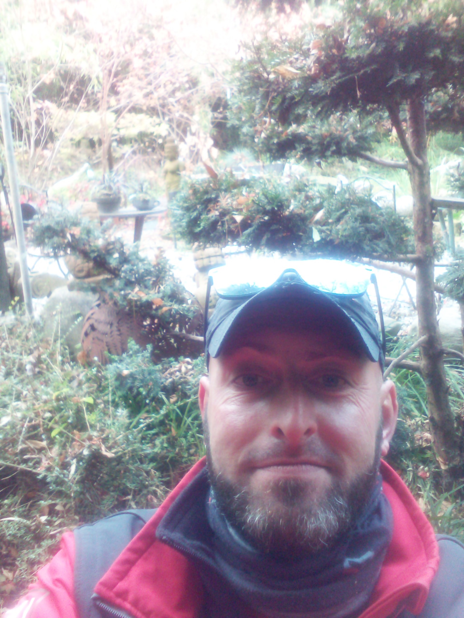 Autoportret mężczyzny w czapce i okularach na tle ogrodu z iglakami i elementami małej architektury.