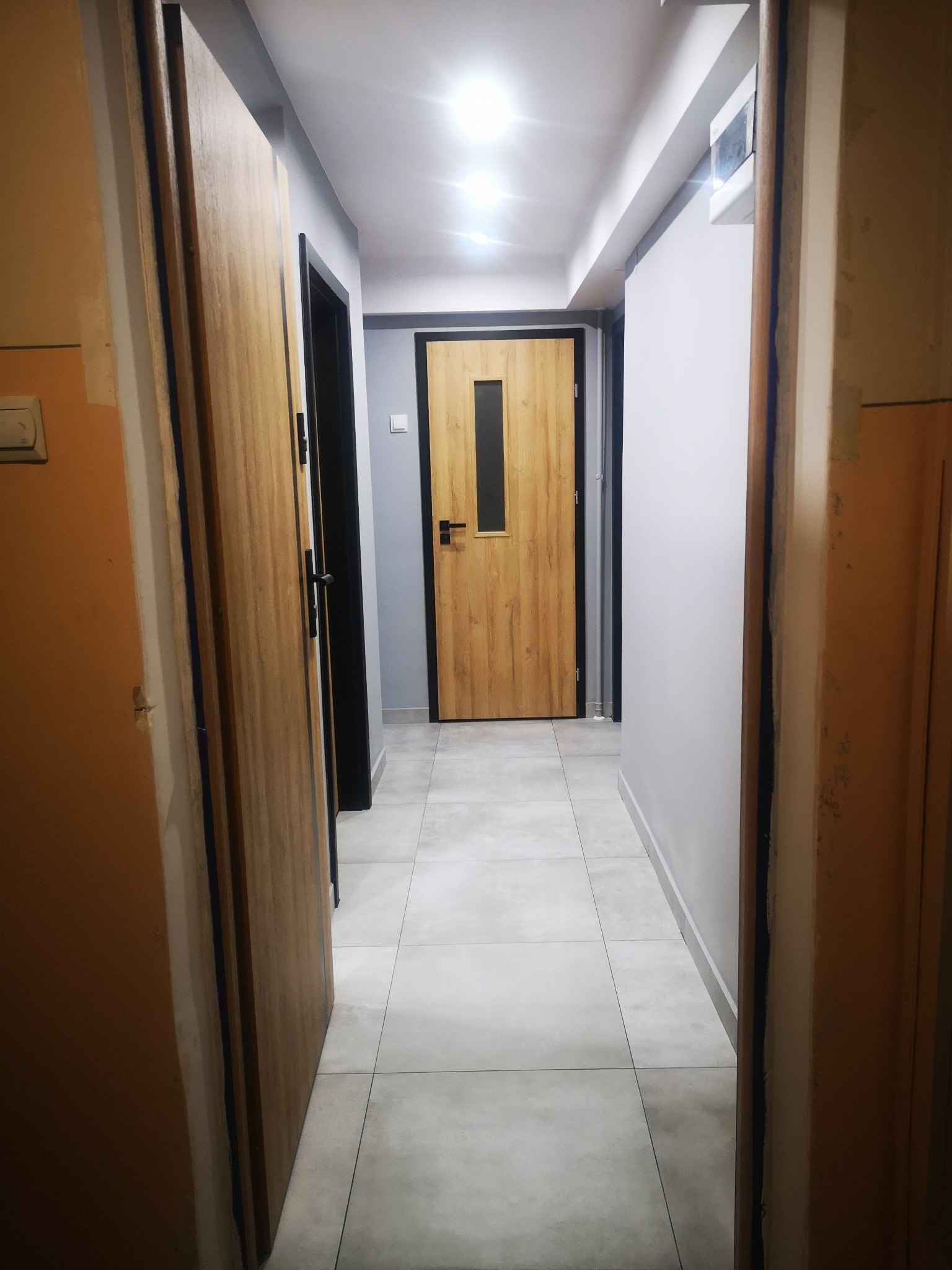 Korytarz po odnowieniu z dwiema drewnianymi ościeżnicami, szarą ścianą i nową drewnianą drzwią z czarną ramą i szybą w głębi.