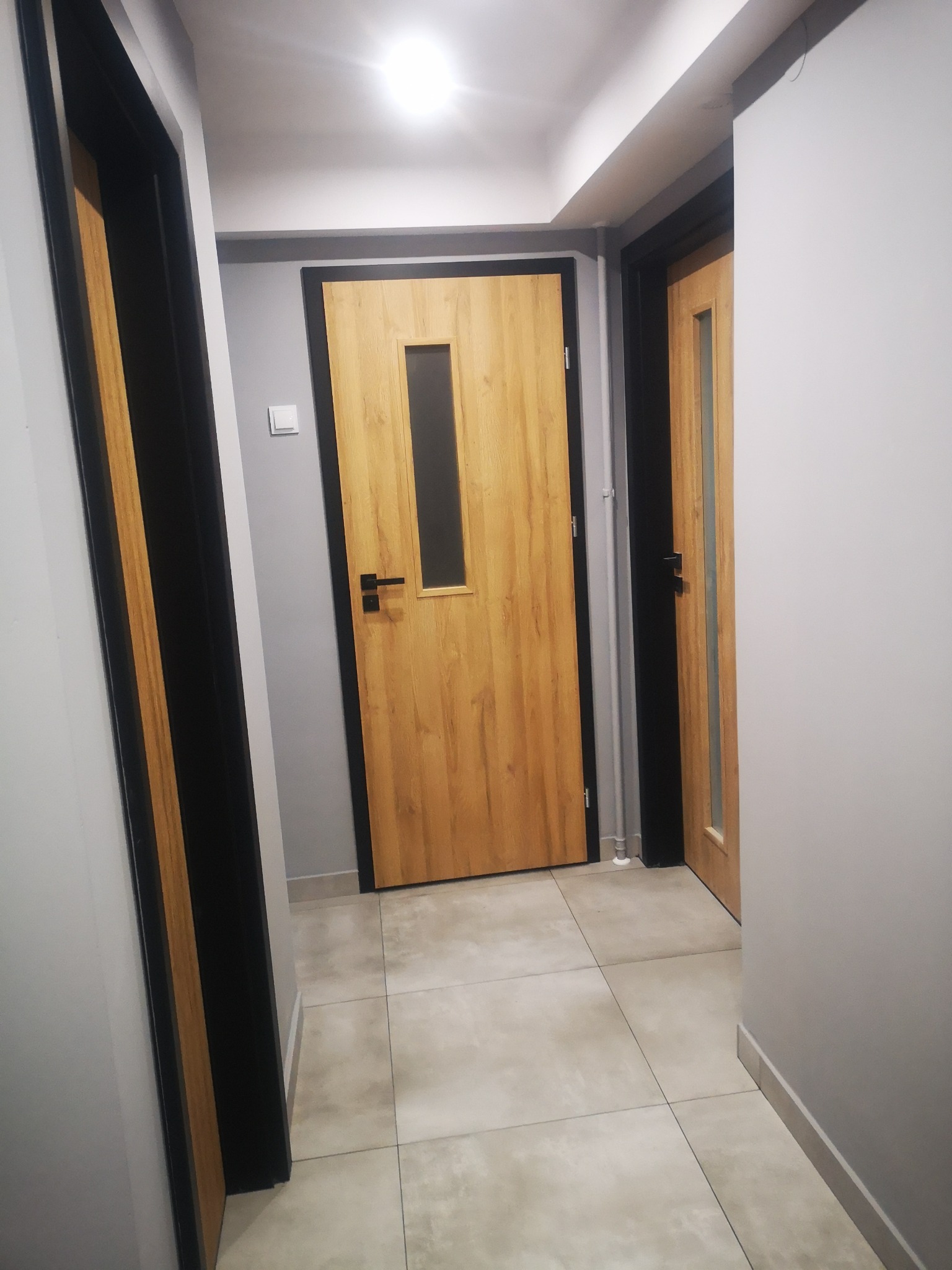 Korytarz z dwiema drewnianymi drzwiami z czarnymi ościeżnicami i przeszkleniami, widok z perspektywy podłogi, szare ściany i płytki podłogowe.