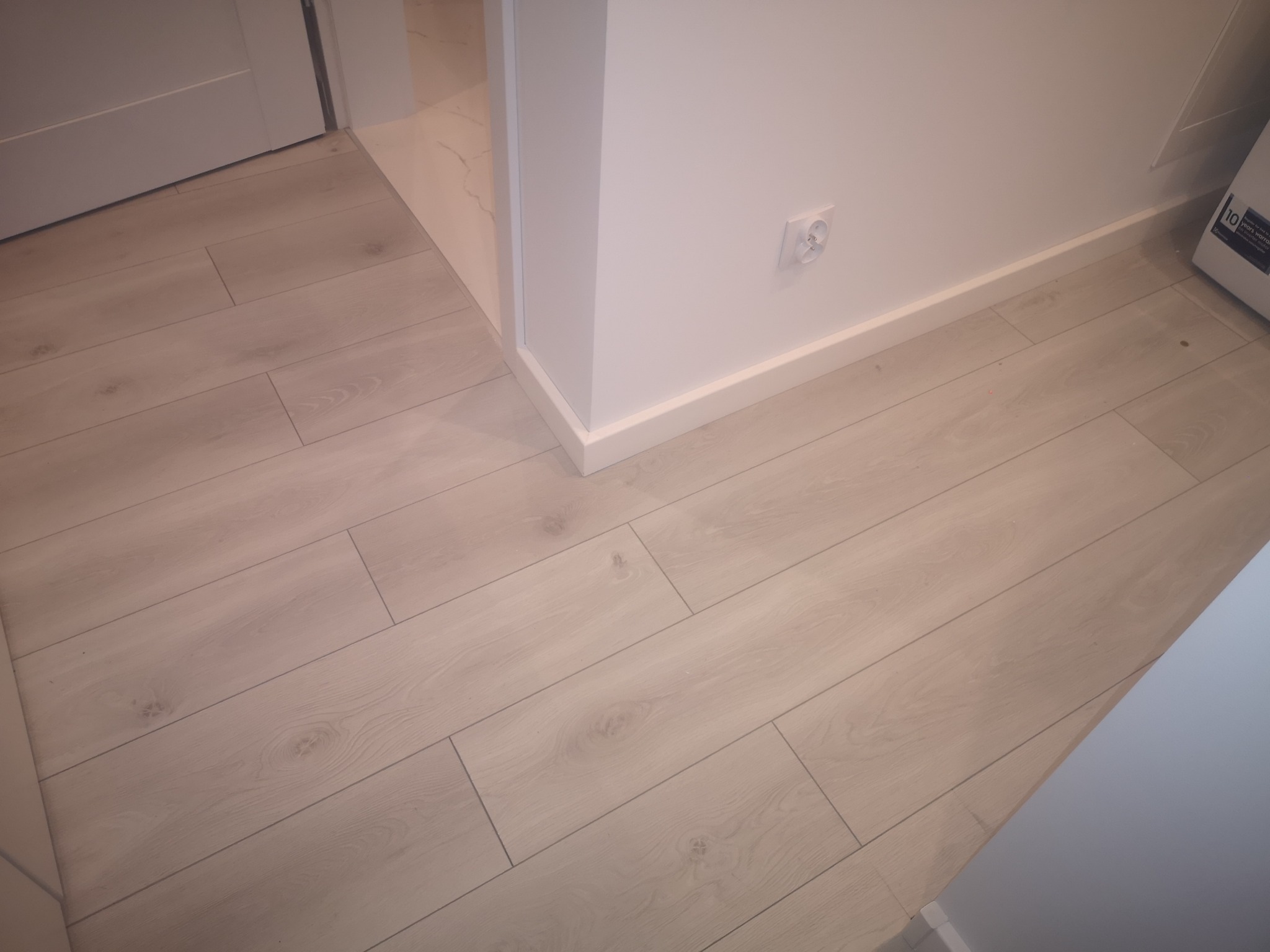 Jasne panele podłogowe ułożone wzdłuż ściany z gniazdkiem, widoczne białe listwy przypodłogowe i fragment drzwi.