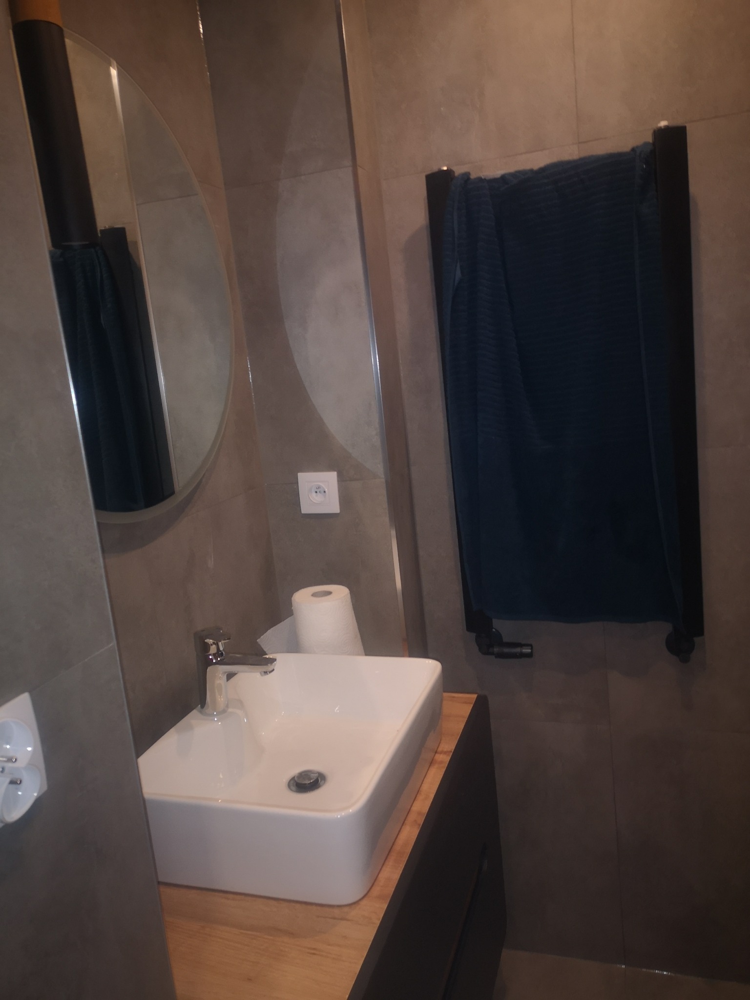 Nowoczesna łazienka z umywalką nablatową na drewnianym blacie, okrągłym lustrem i grzejnikiem drabinkowym z ciemnym ręcznikiem.