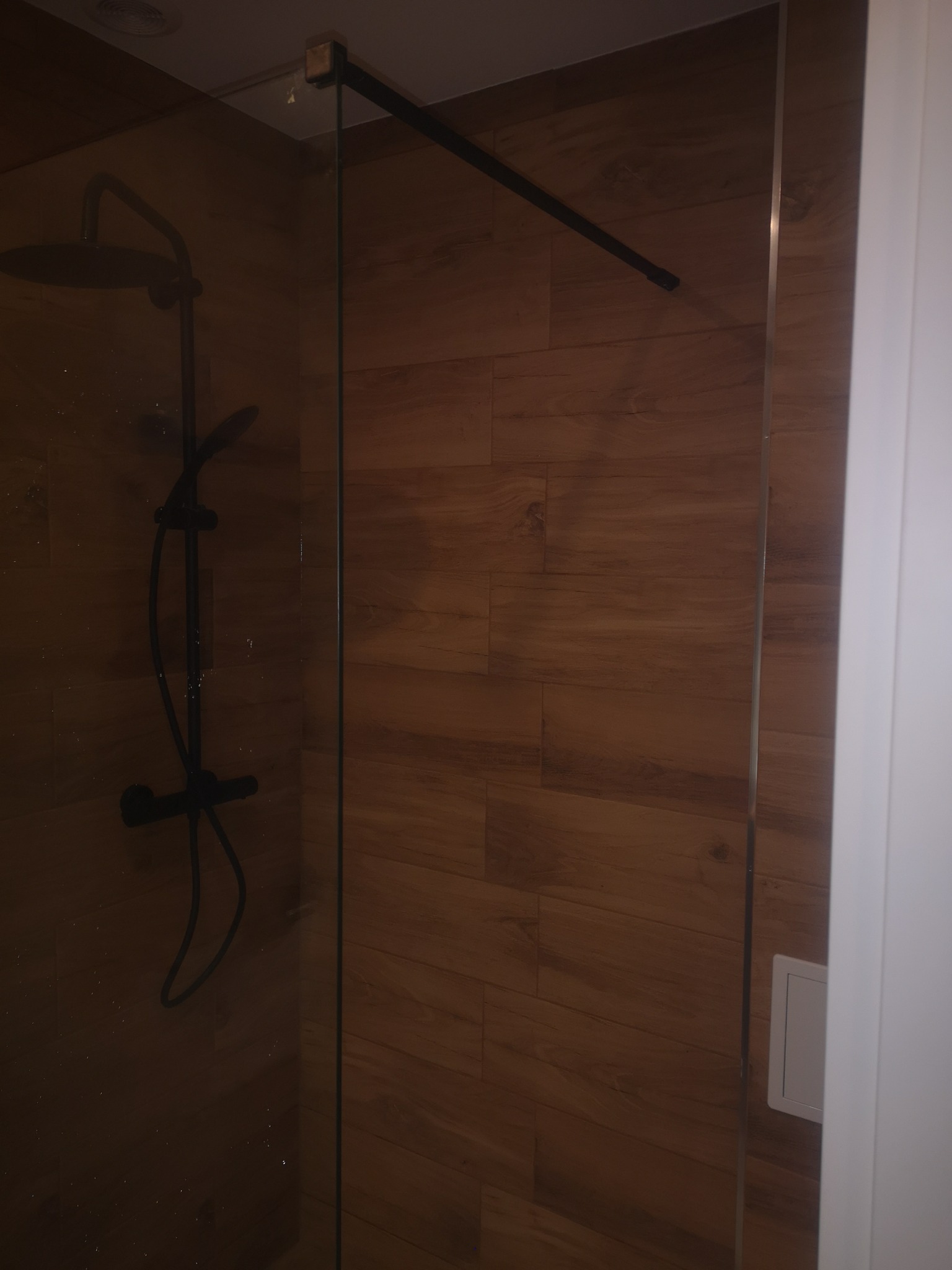 Nowoczesna kabina prysznicowa z czarną armaturą na tle drewnopodobnych płytek ściennych, widok z boku.