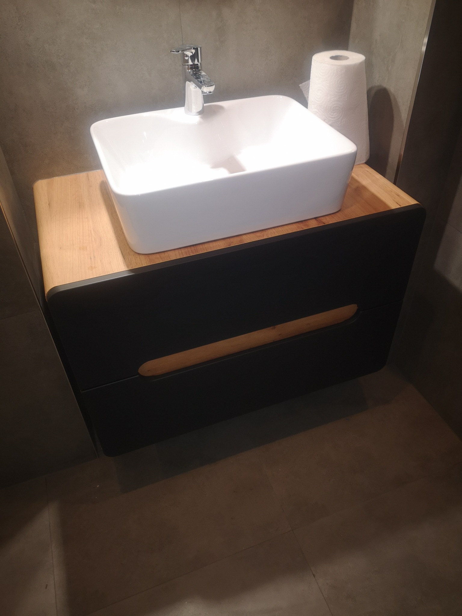 Nowoczesna łazienka: biała, prostokątna umywalka nablatowa z chromowaną baterią, osadzona na szafce z drewnianym blatem i czarnym frontem z podłużnym, zaokrąglonym otworem na uchwyt, obok rolka...