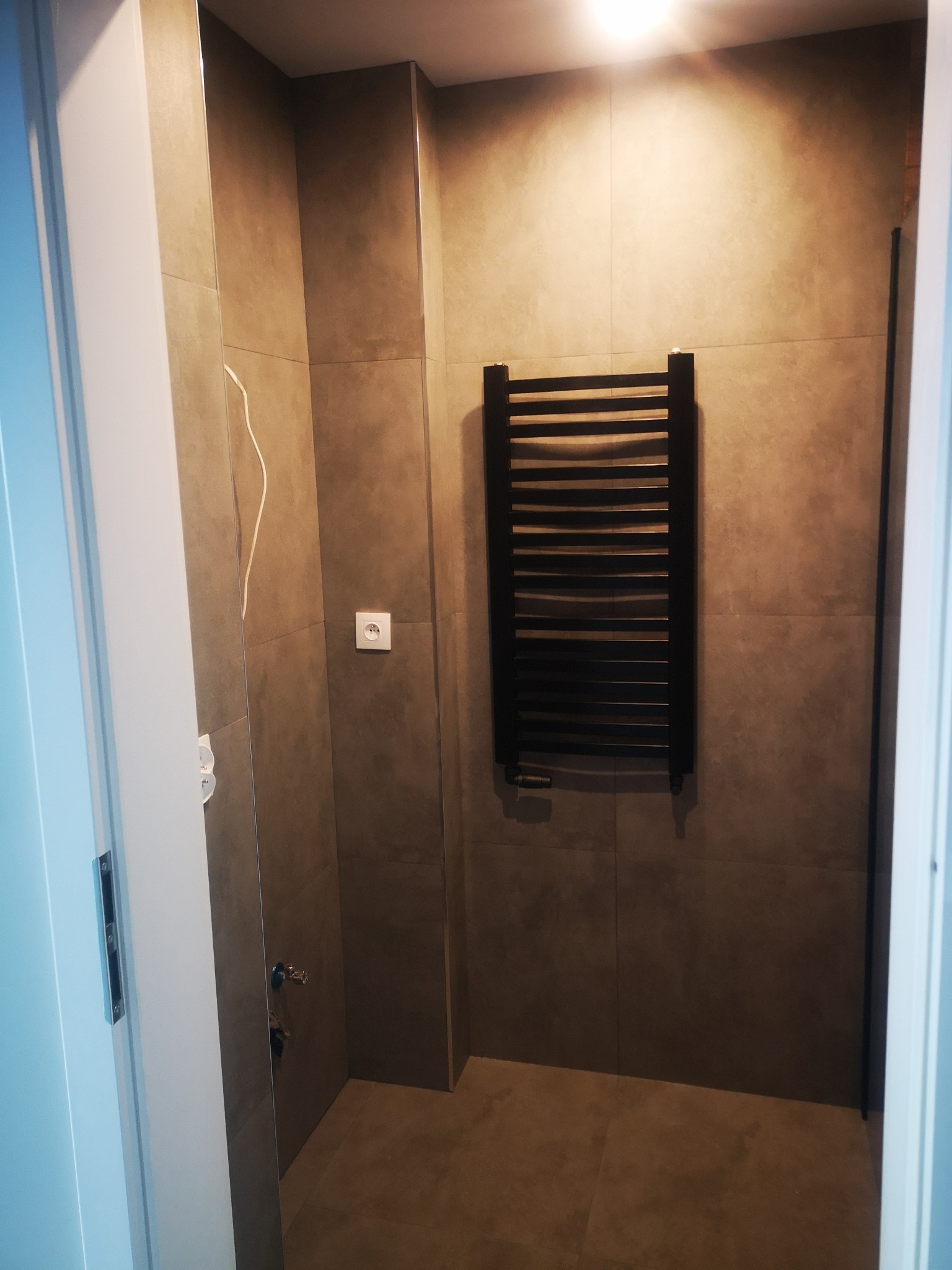 Wykończona łazienka z szarymi płytkami, czarnym grzejnikiem drabinkowym i widocznymi elementami instalacji elektrycznej i wodnej.