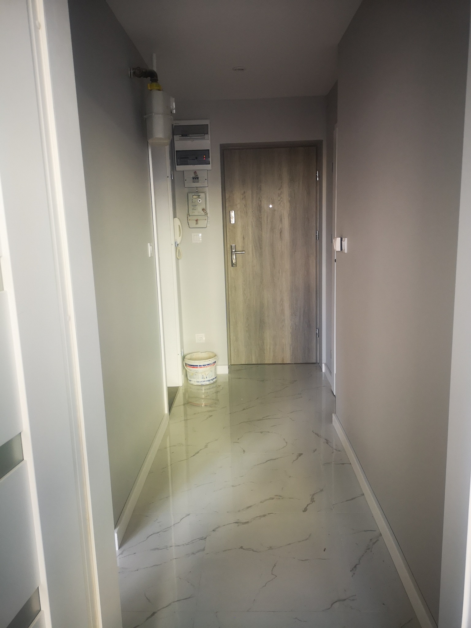 Korytarz po remoncie z szarymi ścianami, białą marmurową podłogą i drewnianymi drzwiami wejściowymi; widoczny licznik gazu i wiadro z farbą w rogu.