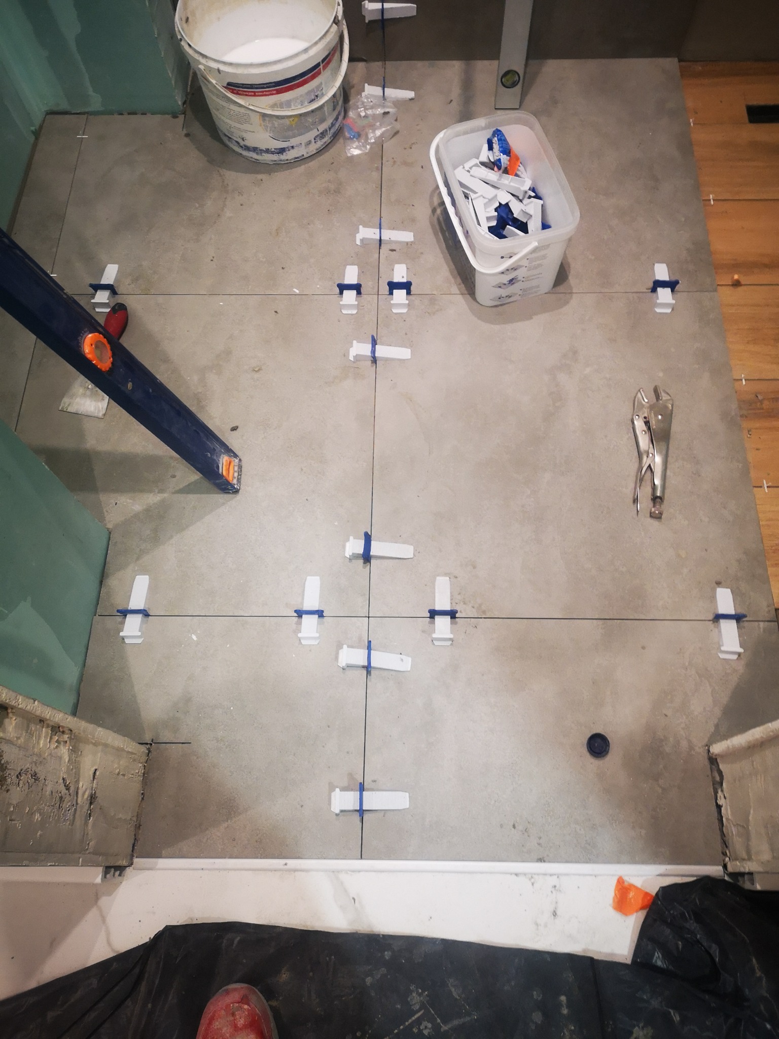 Układanie szarych płytek podłogowych z użyciem systemu poziomowania, widoczny poziomica, wiadro z klejem i szczypce do płytek.