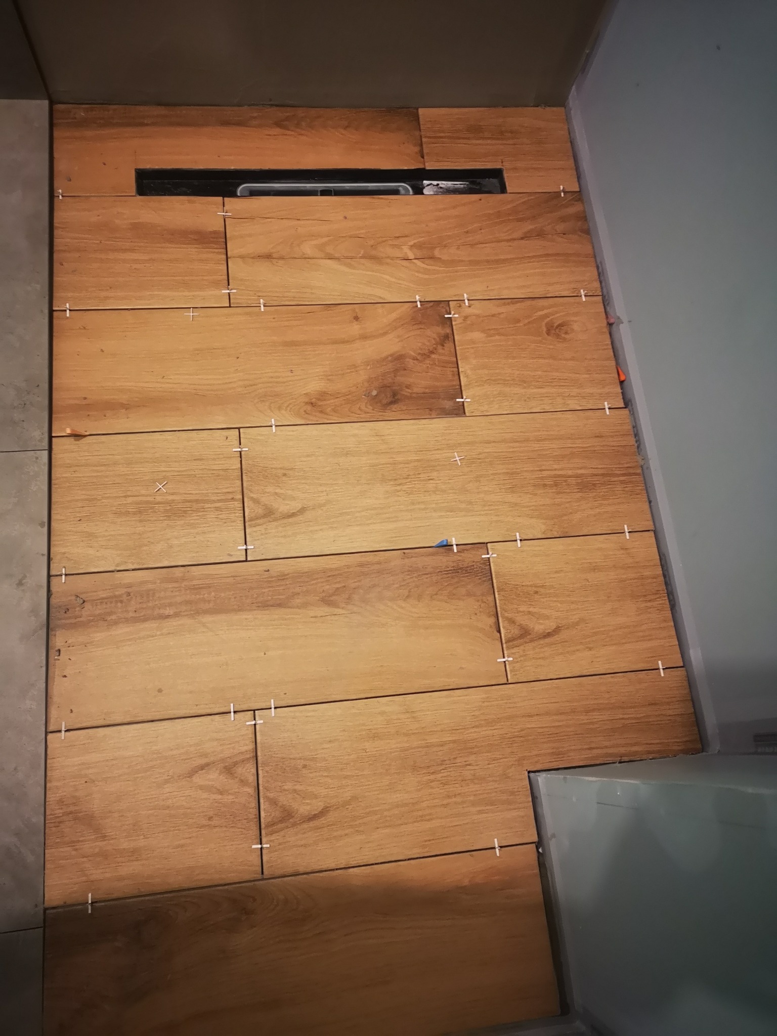 Układanie płytek imitujących drewno na podłodze w łazience, widoczne krzyżyki dystansowe i odpływ liniowy.