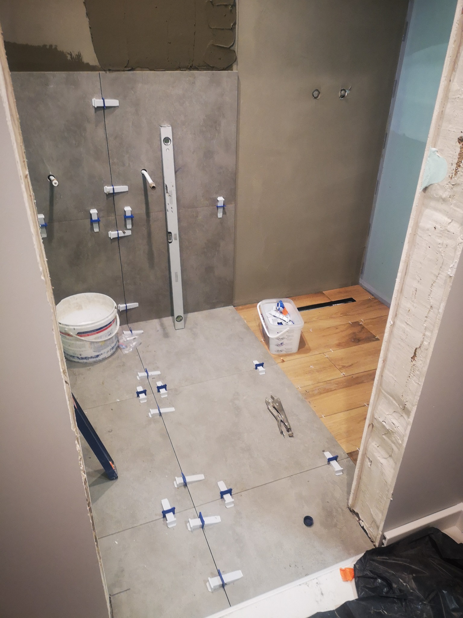 Łazienka w trakcie prac: szare płytki na ścianie i podłodze, system poziomowania płytek, poziomica przy ścianie, fragment drewnianej podłogi w kabinie prysznicowej.