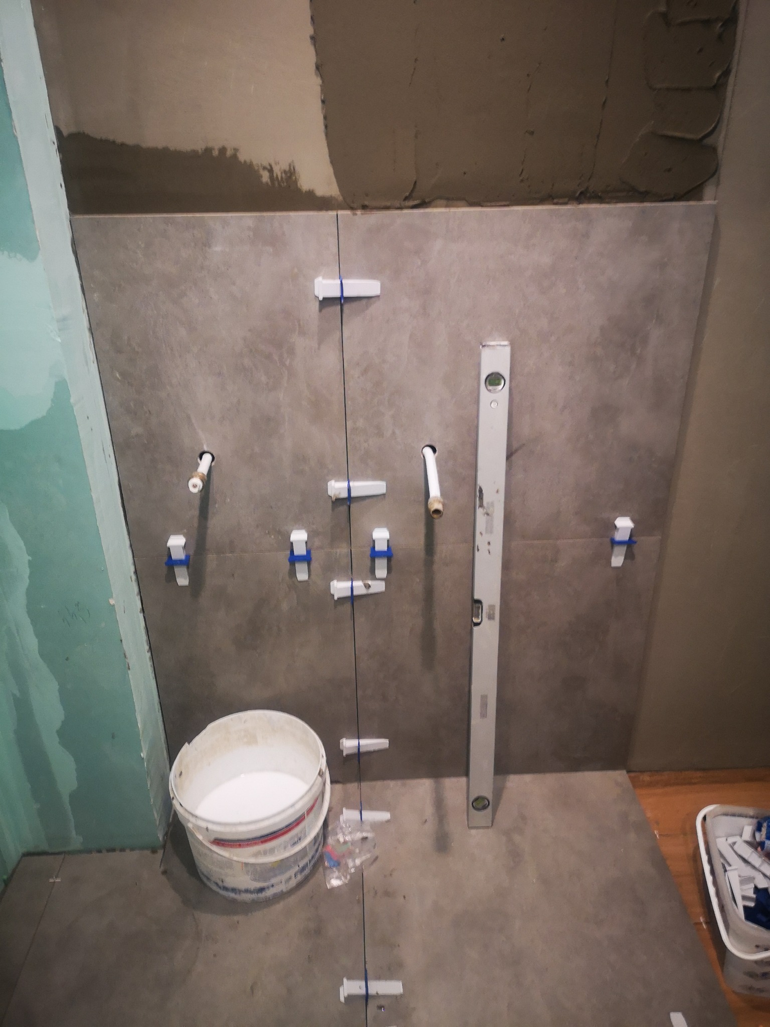 Układanie szarych płytek na ścianie w łazience, widoczne białe krzyżyki dystansowe, poziomica, wiadro z klejem i wystające rurki instalacji wodnej.