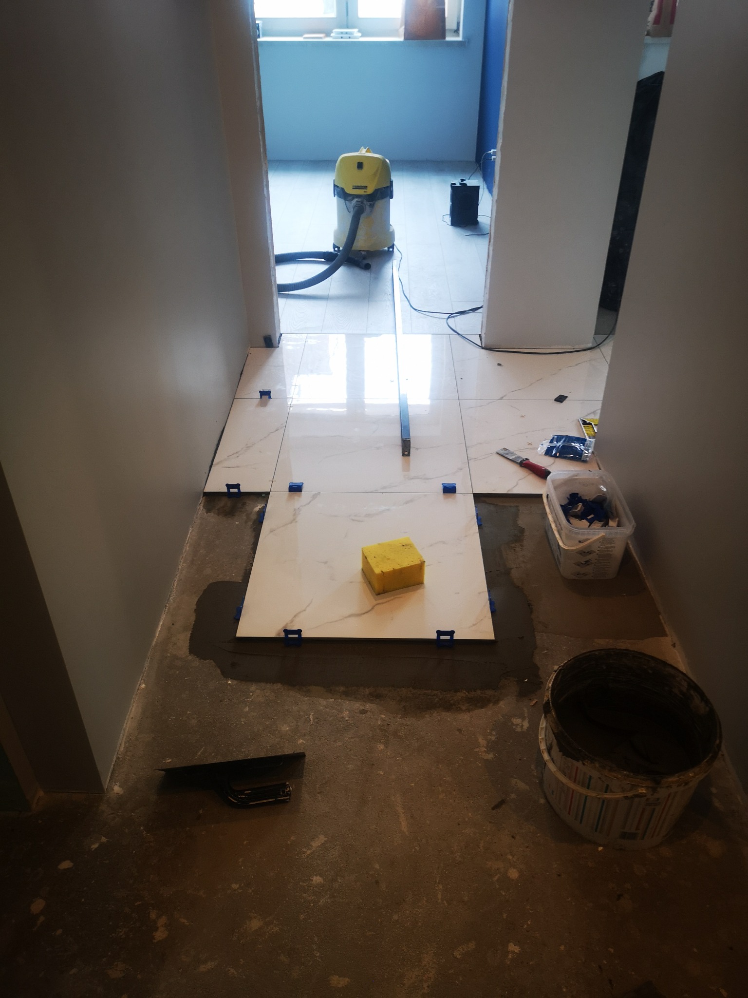 Układanie białych płytek imitujących marmur w korytarzu, widoczny żółty odkurzacz przemysłowy w tle, poziomowanie płytek przy użyciu klipsów i krzyżyków, obok wiadro z zaprawą i paca.