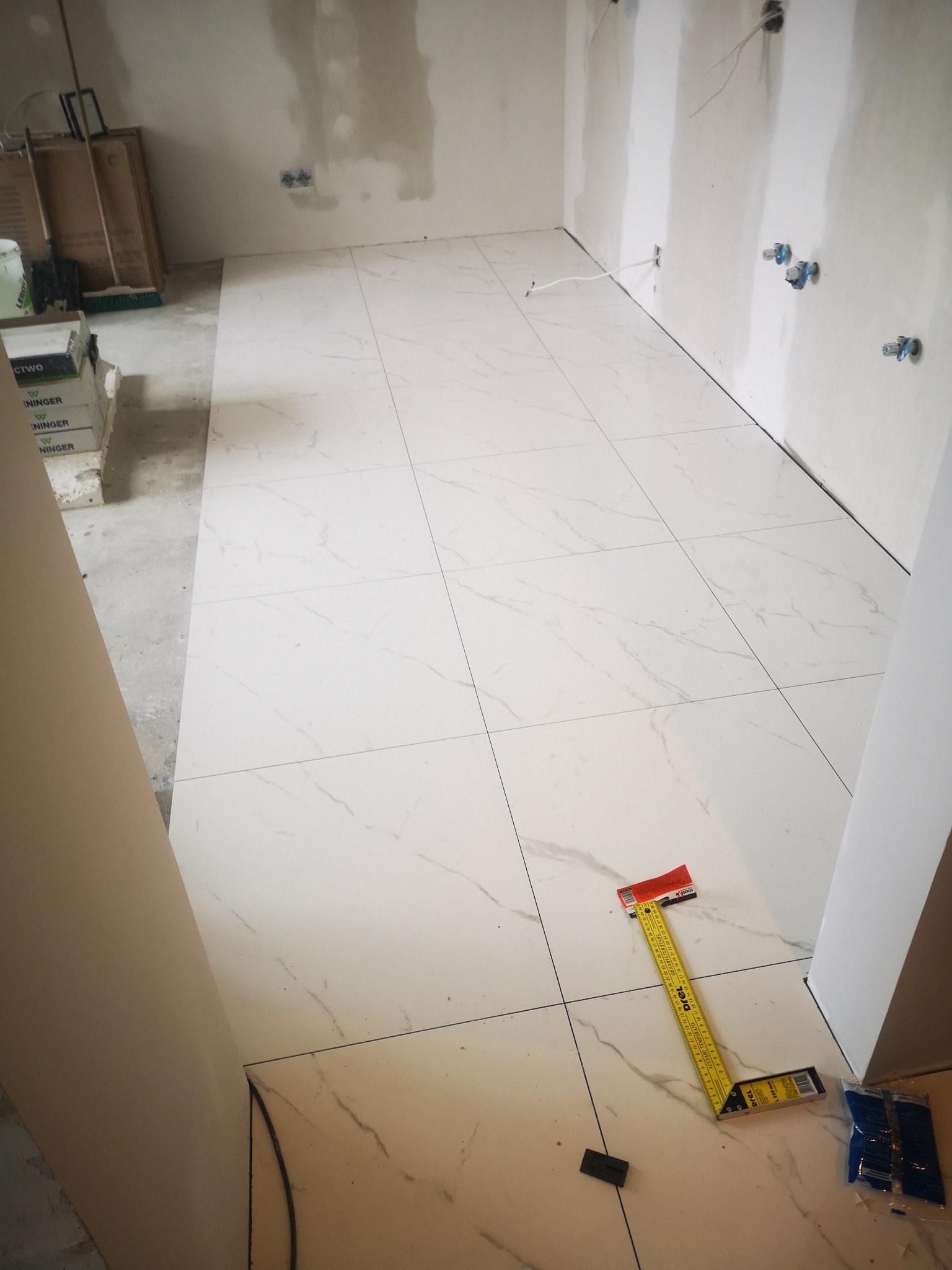 Układanie białych płytek podłogowych z szarym wzorem żyłek w Tarnowie, widoczna poziomnica i krzyżyki dystansowe, ściany przygotowane do dalszych prac wykończeniowych.
