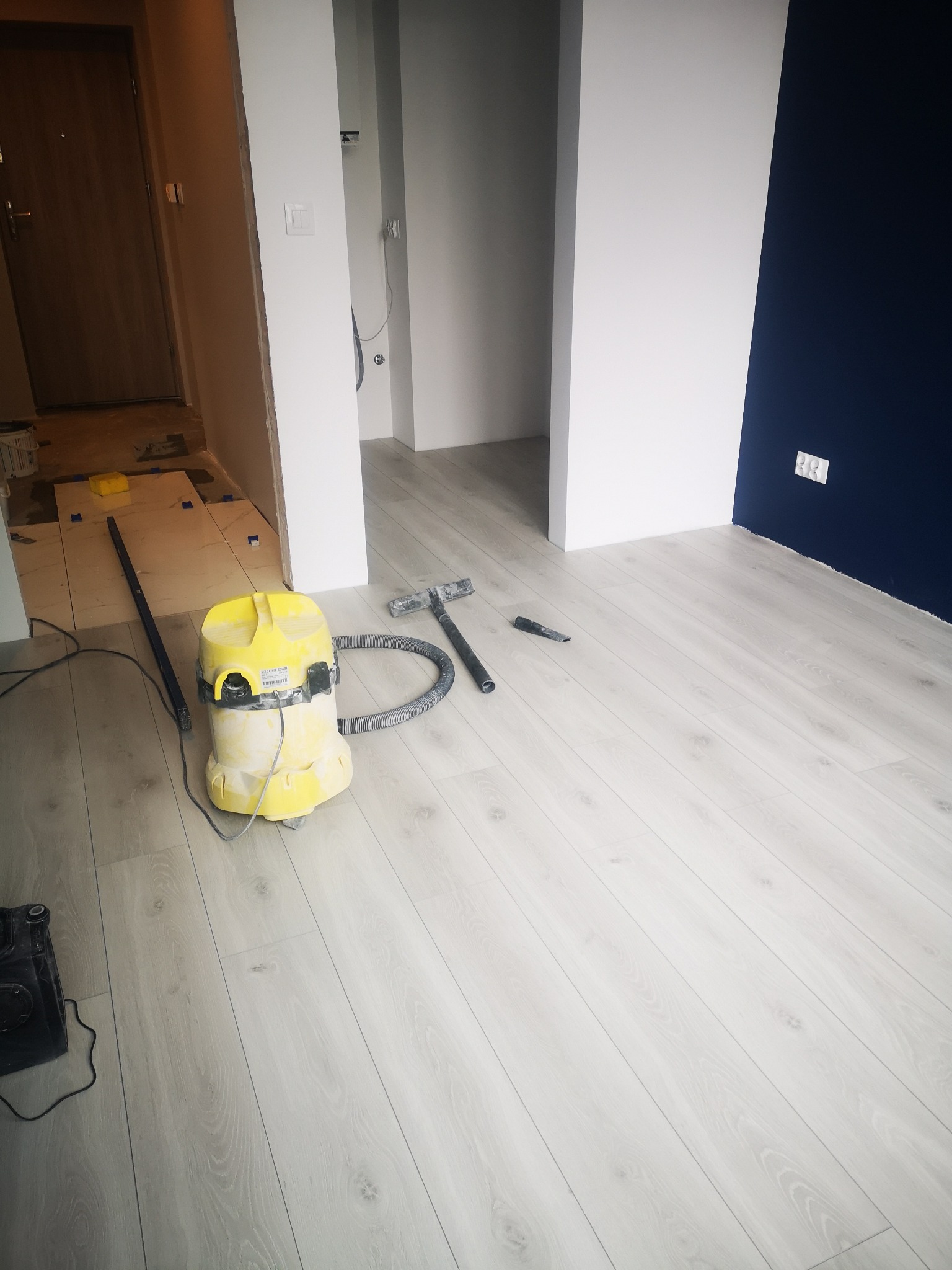 Nowo ułożona, jasna podłoga laminowana w mieszkaniu, widoczny odkurzacz przemysłowy, listwa przypodłogowa do zamontowania i fragment położonych płytek w sąsiednim pomieszczeniu.