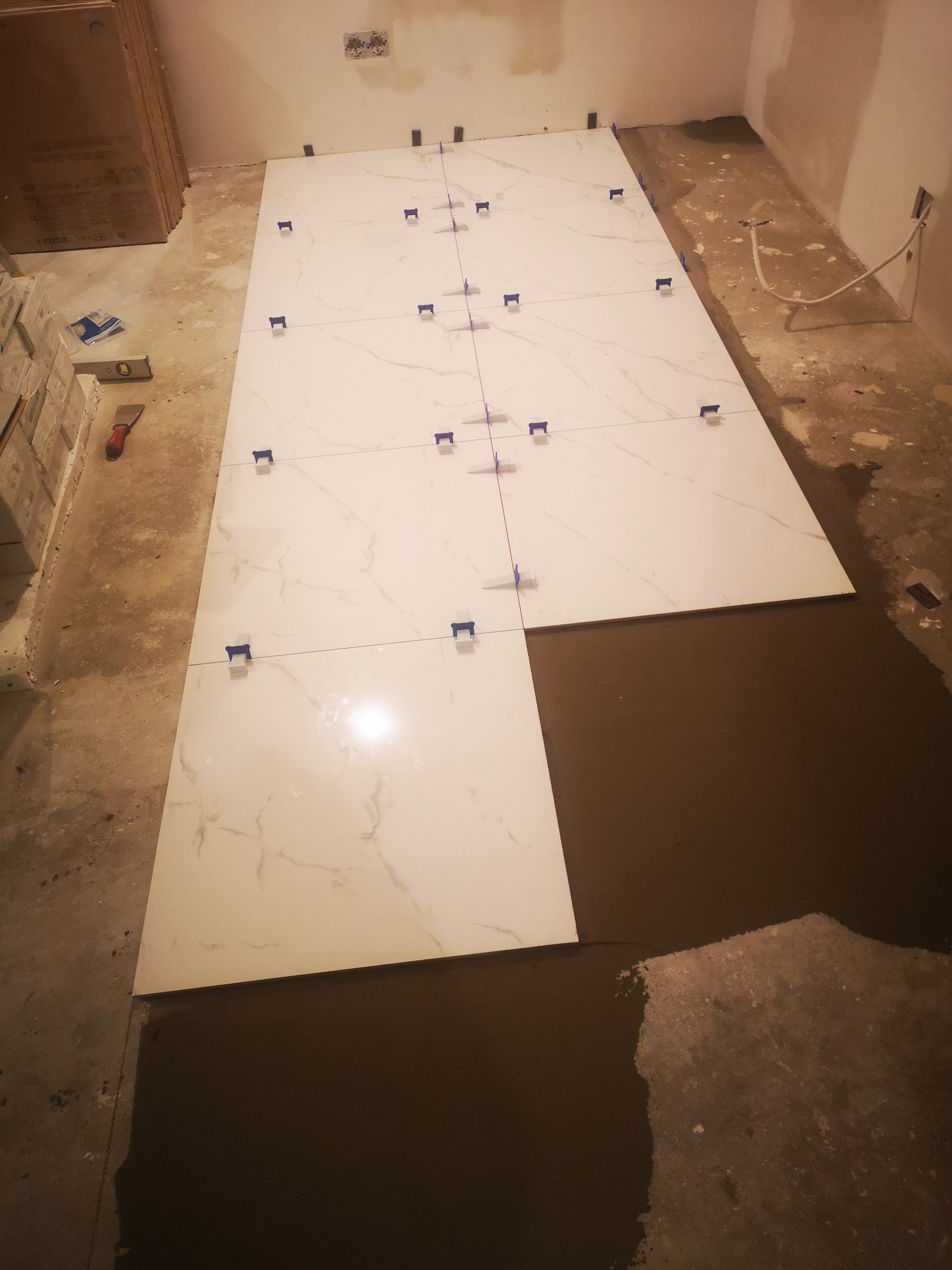 Układanie białych płytek imitujących marmur na podłodze, widoczne niebieskie krzyżyki dystansowe i poziomica, świeżo rozprowadzona zaprawa pod kolejną płytkę.
